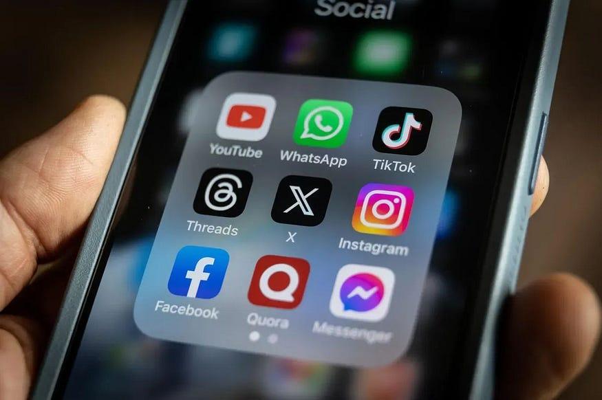 A hand holding a phone showing a folder of social apps — YouTube, WhatsApp, TikTok, Threads, X, Instagram, Facebook, Quora and Messenger