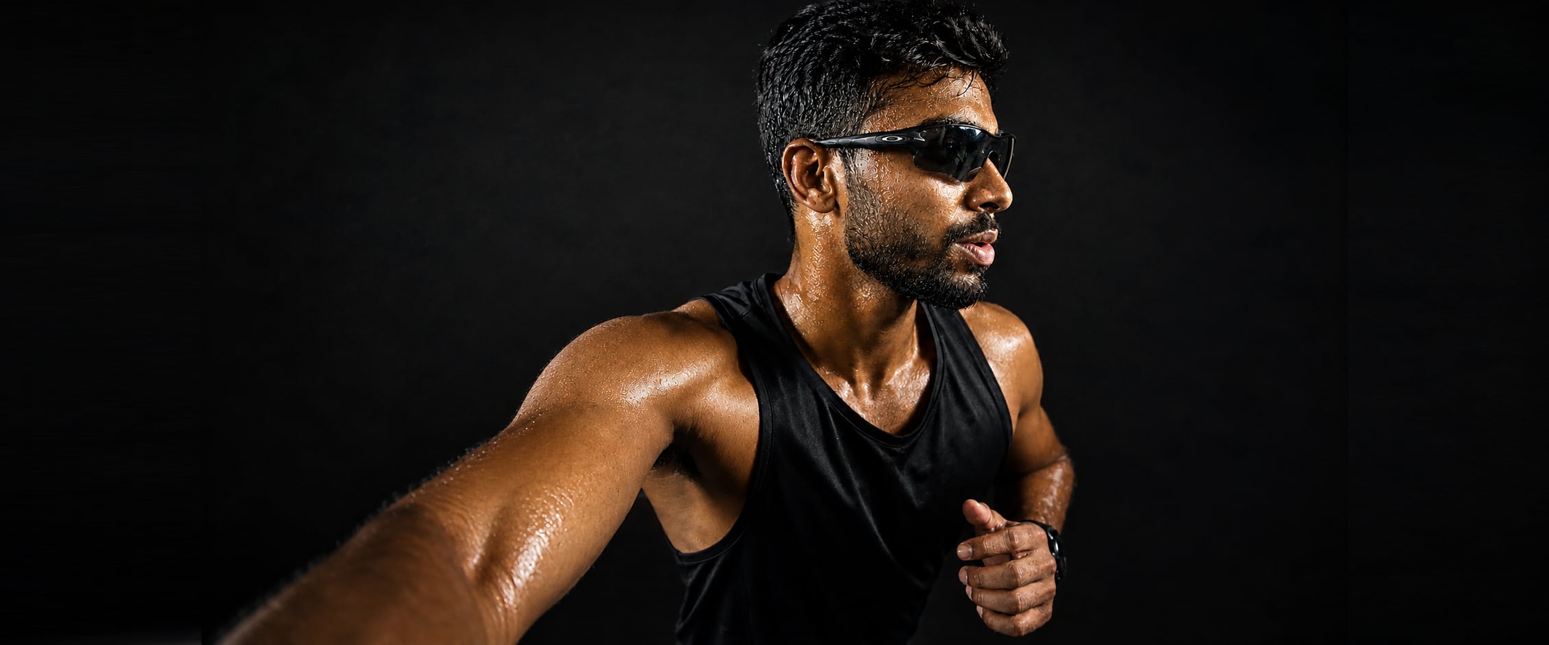 A sweating athlete in sunglasses and a black vest, mid-stride against a dark background.
