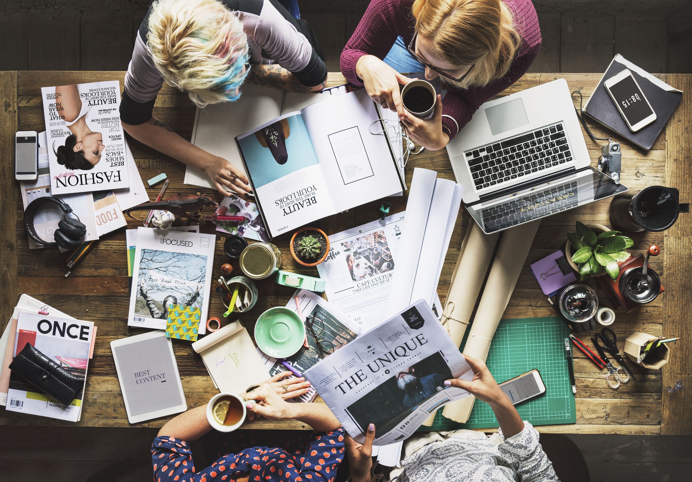 An overhead view of colleagues collaborating around a desk covered with magazines, laptops and coffee.